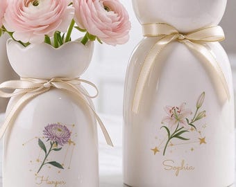 May include: Two white ceramic vases with scalloped rims and cream-coloured ribbons. The smaller vase holds pink ranunculus and features a purple flower illustration with the name Harper. The larger vase holds white eustoma and a lily illustration with the name Sophia.