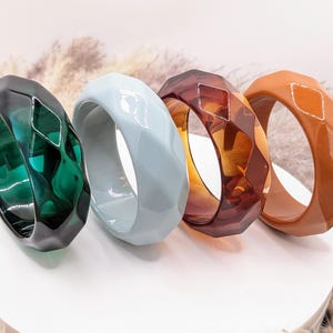 May include: Four faceted bangle bracelets arranged in a row. The first is translucent emerald green, the second is opaque light blue, the third is translucent dark amber, and the fourth is opaque terracotta orange. Each bracelet has a geometric design.