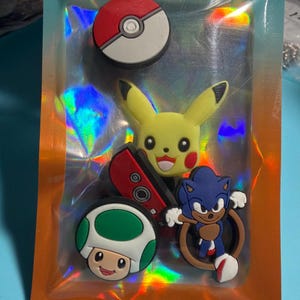 May include: A set of five colorful rubber shoe charms featuring a red and white Poké Ball, a yellow Pikachu character, a red game controller, a green and white mushroom character, and a blue Sonic the Hedgehog character inside a holographic pouch.