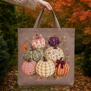 May include: A taupe canvas tote bag featuring a central illustration of seven pumpkins in various colors and patterns, including plaid, floral, and scalloped designs, accented with small stars, swirling vines, and a dark red ribbon bow.