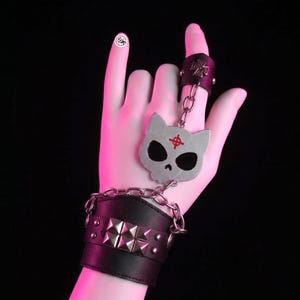 Peut inclure: Un bracelet de poignet en cuir noir orné d'une rangée de clous pyramidaux argentés, relié par une chaîne en argent à un pendentif en métal en forme de crâne de chat avec un symbole de croix rouge, et une bague assortie avec une croix en relief.