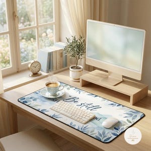May include: A rectangular desk mat featuring a white background with blue and green watercolor-style leaf patterns along the edges. The center of the mat displays the text "Be still" in a dark blue cursive script font, providing a workspace surface.
