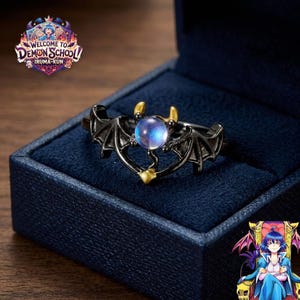 May include: A black metal ring featuring a central iridescent blue orb flanked by two bat-like wings and two small gold-colored horns. The ring is presented in a dark blue velvet-lined box, with the Welcome to Demon School Iruma-kun logo visible.