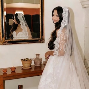 May include: A white wedding veil featuring intricate lace trim along the edges. The veil is embroidered with the white cursive text Mrs. Smith and the date October 26, 2024. The sheer fabric drapes over the shoulders, showcasing detailed floral lace patterns.