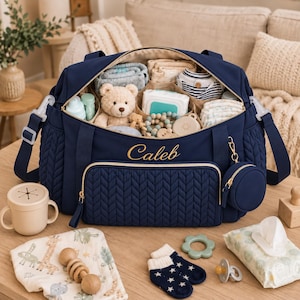 Pode incluir: Uma mala de maternidade acolchoada azul-marinho com o nome Caleb bordado a dourado no bolso frontal. A mala está aberta, revelando artigos essenciais para bebé organizados, incluindo um urso de peluche, toalhitas e roupa. Inclui uma bolsa.