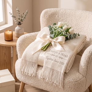 Puede incluir: Una manta texturizada de color crema con flecos, bordada con el texto "Evelyn + Noah" y "Together Since 2026" junto a una línea decorativa y un corazón. La manta está envuelta con una cinta de satén color crema y decorada con tulipanes blancos.