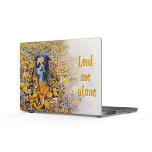 May include: A laptop skin featuring a photograph of a dark-furred dog sitting in a pile of vibrant yellow and orange autumn leaves with a single leaf on its head. The text "Leaf me alone" is printed in a gold-toned font on the right side.