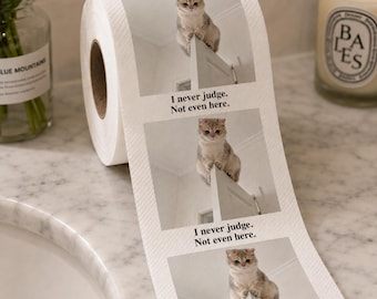 Könnte beinhalten: Eine Rolle weißes Toilettenpapier mit einem wiederholten Fotodruck einer hellbraunen Katze, die auf einer Tür sitzt. Unter jedem Bild der Katze ist der Text "I never judge. Not even here." in einer einfachen schwarzen serifenlosen Schrift gedruckt.