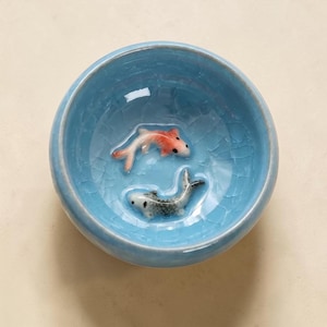 May include: A round ceramic bowl featuring a light blue crackle glaze finish. Inside the bowl, two small, three-dimensional fish figures are attached to the bottom. One fish is orange and white, while the other is grey and white with black spots.