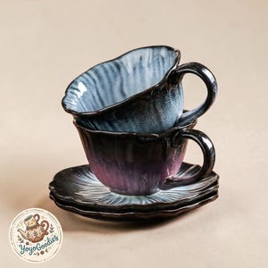 Puede incluir: Dos tazas de té de cerámica apiladas con platillos a juego que presentan un borde ondulado y festoneado. La taza superior tiene un interior azul claro y blanco, mientras que la taza inferior y los platillos muestran un degradado morado y negro.