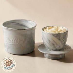 Op de afbeelding: Een keramische boterpot met een grijs en wit marmerpatroon. De basiscontainer heeft het woord Butter in reliëf op de zijkant. Het deksel ligt omgekeerd naast de basis, gevuld met een krul gele boter, en toont de marmeren afwerking.