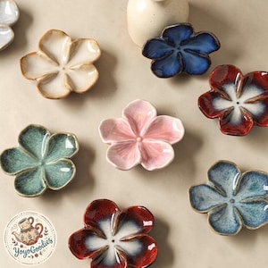 Puede incluir: Una colección de platos de cerámica con forma de flor en varios colores, incluyendo crema, azul, rojo, rosa y verde azulado. Cada plato presenta cinco pétalos con un acabado brillante. Un logotipo circular con una ilustración de una tetera y el texto YoyoGoodies es visible.
