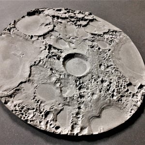 Puede incluir: Un disco circular gris que presenta un mapa en relieve tridimensional texturizado de la superficie lunar. El objeto muestra numerosos cráteres, depresiones y terreno irregular en toda su cara superior, imitando la apariencia de la luna.