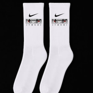 May include: A pair of white crew socks featuring a black Nike swoosh logo at the top. Below the logo is a rectangular graphic showing a pair of intense red eyes with black markings, and the text ITACHI printed in black capital letters underneath.
