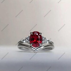 Könnte beinhalten: Ein silberfarbener Ring mit einem zentralen, oval geschliffenen, tiefroten Edelstein, der von vier Krappen gehalten wird. Die Ringschiene hat eine V-Form und ist auf beiden Seiten des Mittelsteins mit je drei kleinen, klaren, runden weißen Steinen besetzt.