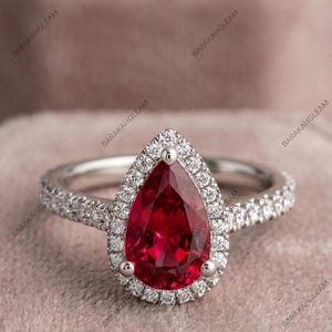 Könnte beinhalten: Ein silberfarbener Ring mit einem birnenförmigen, tiefroten Edelstein als Mittelstück. Der Stein ist von einem Heiligenschein aus kleinen, klaren, rund geschliffenen Steinen umgeben. Die Ringschiene ist ebenfalls mit kleinen, klaren Steinen besetzt.