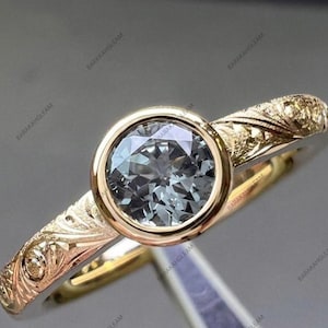 Könnte beinhalten: Ein goldener Ring mit einem runden, facettierten, grauen Mittelstein in einer glatten Zargenfassung. Die Ringschiene besteht aus Gold und weist entlang des Schafts komplizierte, blumenartige Gravuren auf, die von der Fassung ausgehen.
