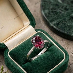 Könnte beinhalten: Ein silberner Ring mit einem großen, oval geschliffenen, tiefroten Edelstein, der von vier Krappen gehalten wird. Der Ring befindet sich in einer geöffneten, dunkelgrünen Schmuckschatulle aus Samt mit cremefarbenem Innenfutter.
