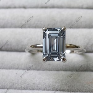 Könnte beinhalten: Ein silberfarbener Ring mit einem großen, rechteckigen Stein im Smaragdschliff in einem kühlen Graublau. Der Edelstein wird von vier Metallkrappen auf einer glatten, polierten Ringschiene gehalten. Das Design ist mittig und symmetrisch.