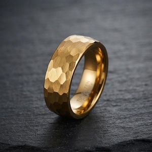 May include: A gold-coloured ring with a hammered, faceted exterior texture. The interior of the band is smooth and features the engraved text Tungsten Carbide. The ring has a wide, flat profile and a polished finish that reflects light across its surface.