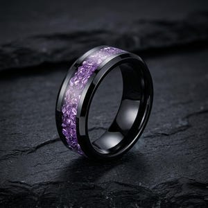 Peut inclure: Une bague noire dotée d'une incrustation centrale de fragments de pierre violette concassée. L'anneau présente une finition lisse et polie avec des bords biseautés, créant un contraste entre la surface métallique sombre et l'incrustation texturée.