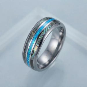 Puede incluir: Un anillo de tono plateado pulido con una incrustación central de ópalo azul brillante flanqueada por dos bandas de concha de abulón iridiscente. La banda tiene un perfil exterior liso y redondeado con un acabado metálico reflectante.