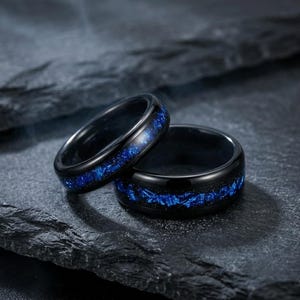 Puede incluir: Dos anillos negros con una incrustación central de material azul triturado que brilla con la luz. Las bandas tienen un acabado liso y pulido con un perfil redondeado, mostrando un profundo contraste oscuro frente a los vibrantes acentos azules.