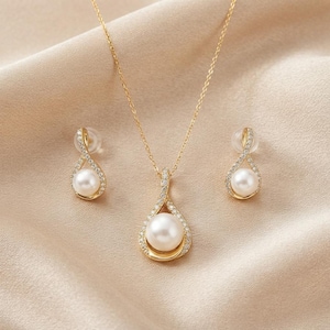 Puede incluir: Un conjunto de joyería dorada que consta de un collar con colgante y dos pendientes a juego. Cada pieza presenta una perla blanca engastada en un marco dorado en forma de lágrima, decorado con pequeñas piedras claras en los bordes.