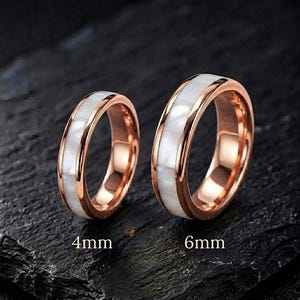 May include: Two rose gold rings featuring a central inlay of white iridescent shell segments. The left ring is labeled 4mm and the right ring is labeled 6mm, indicating their respective widths. Both bands have a polished metallic finish.
