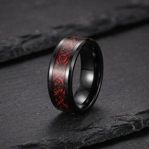 May include: A black ring featuring a polished finish and a central inlay with a vibrant red background. The inlay is decorated with a continuous, intricate black Celtic knotwork pattern that wraps around the entire circumference of the band.
