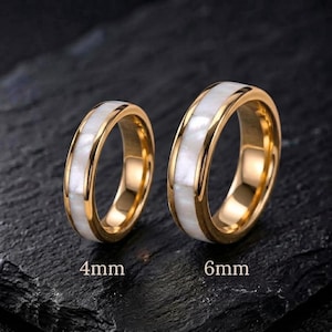 May include: Two gold-toned rings featuring a central inlay of white iridescent shell material. The ring on the left is labeled 4mm and the ring on the right is labeled 6mm, indicating their respective widths. Both bands have a polished finish.