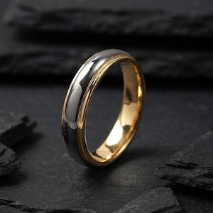 Könnte beinhalten: Ein zweifarbiger Metallring mit einem polierten silberfarbenen Außenband und einem glatten goldfarbenen Innenband. Der Ring hat ein abgerundetes Profil mit einer reflektierenden Oberfläche, die einen klaren Kontrast zwischen den Farben zeigt.