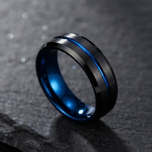 Op de afbeelding: Een zwarte ring met een geborstelde afwerking en een dunne, levendige blauwe groef die horizontaal rond het midden loopt. De binnenkant van de band is gepolijst en heeft een helderblauwe afwerking die contrasteert met het donkere oppervlak.