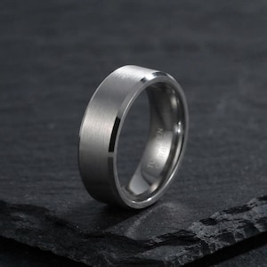 Puede incluir: Un anillo de carburo de tungsteno de tono plateado con un acabado mate cepillado y bordes biselados pulidos. El interior de la banda es liso y reflectante, con el texto TUNGSTEN CARBIDE grabado con láser en la superficie interna.
