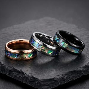 Op de afbeelding: Drie ringen met gefacetteerde randen en inlays van iriserende abalone-schelp. Eén ring heeft een roségouden tint, één een zilveren tint en één een zwarte tint. Elke band heeft een gladde binnenkant en een textuurrijk, veelkleurig midden.