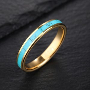 May include: A gold-toned metal ring featuring a central inlay of bright turquoise stone with dark veining. The band has a smooth, polished finish and a rounded profile, showcasing a vibrant contrast between the metallic gold and the blue stone.