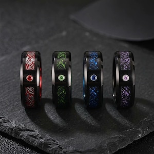 May include: Four black metal rings featuring a central inlay with intricate patterns in red, green, blue, and purple. Each ring has a single round gemstone set in the center of the band, matching the color of the inlay pattern behind it.