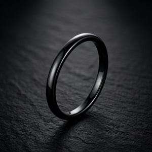 Puede incluir: Un anillo negro sólido con un acabado liso y pulido y un perfil redondeado. La banda presenta un diseño continuo y sin costuras con una superficie de alto brillo que refleja la luz, creando una apariencia elegante y uniforme en toda la pieza.