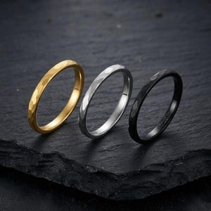 May include: Three thin rings with a hammered texture, arranged in a row. The first ring is gold-toned, the second is silver-toned, and the third is black with the text Tungsten Carbide engraved on the inner surface of the band.