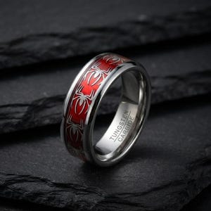 Puede incluir: Un anillo de carburo de tungsteno en tono plateado con una banda incrustada de color rojo vibrante decorada con un patrón repetitivo de siluetas de arañas plateadas. El interior tiene grabado el texto TUNGSTEN CARBIDE en mayúsculas.