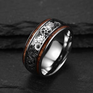 Puede incluir: Un anillo de metal plateado con una incrustación central de madera oscura y una banda negra decorada con un patrón repetitivo de varias formas de engranajes mecánicos. El interior del anillo es pulido y liso con un acabado metálico brillante.