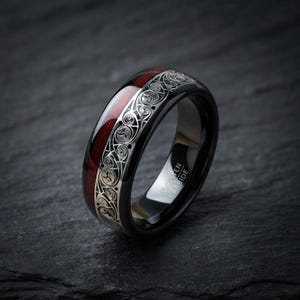 May include: A black tungsten carbide ring featuring a polished finish, a dark red wood inlay, and a silver-toned band engraved with intricate swirling scrollwork patterns. The interior of the band is clearly marked with the text Tungsten Carbide.