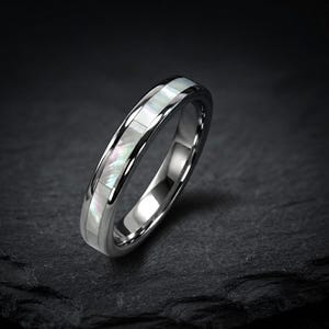 Puede incluir: Un anillo de metal plateado pulido con una incrustación central de segmentos de nácar blanco iridiscente. La banda tiene un perfil liso y redondeado con un acabado reflectante que resalta el brillo nacarado de la incrustación.