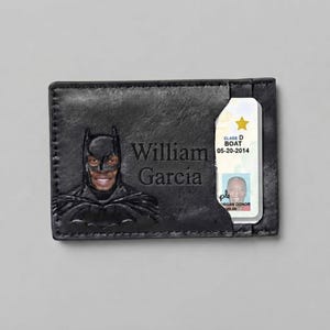 Puede incluir: Un tarjetero de cuero negro con una máscara de Batman en relieve y el rostro de una persona visible en el recorte. El nombre William Garcia está grabado en el frente. Se ha insertado una licencia de conducir con una estrella dorada y el texto Class D BOAT 05-20-2014.