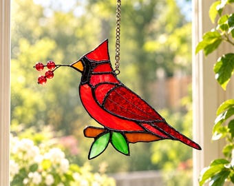 May include: A stained glass suncatcher shaped like a red cardinal bird perched on a branch with green leaves. The bird holds a small stem with three red glass berries in its beak. It is suspended by a metal chain against a window.