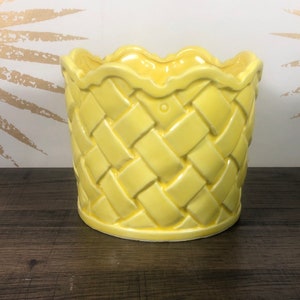 Op de afbeelding: Een gele keramische plantenpot met een geweven patroon. De pot heeft een gegolfde rand en een afgeronde basis.