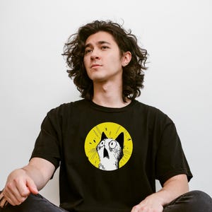 May include: A black short-sleeved t-shirt featuring a circular graphic on the chest. The graphic depicts a white cat with wide, surprised eyes and an open mouth, set against a bright yellow background with black radiating lines.