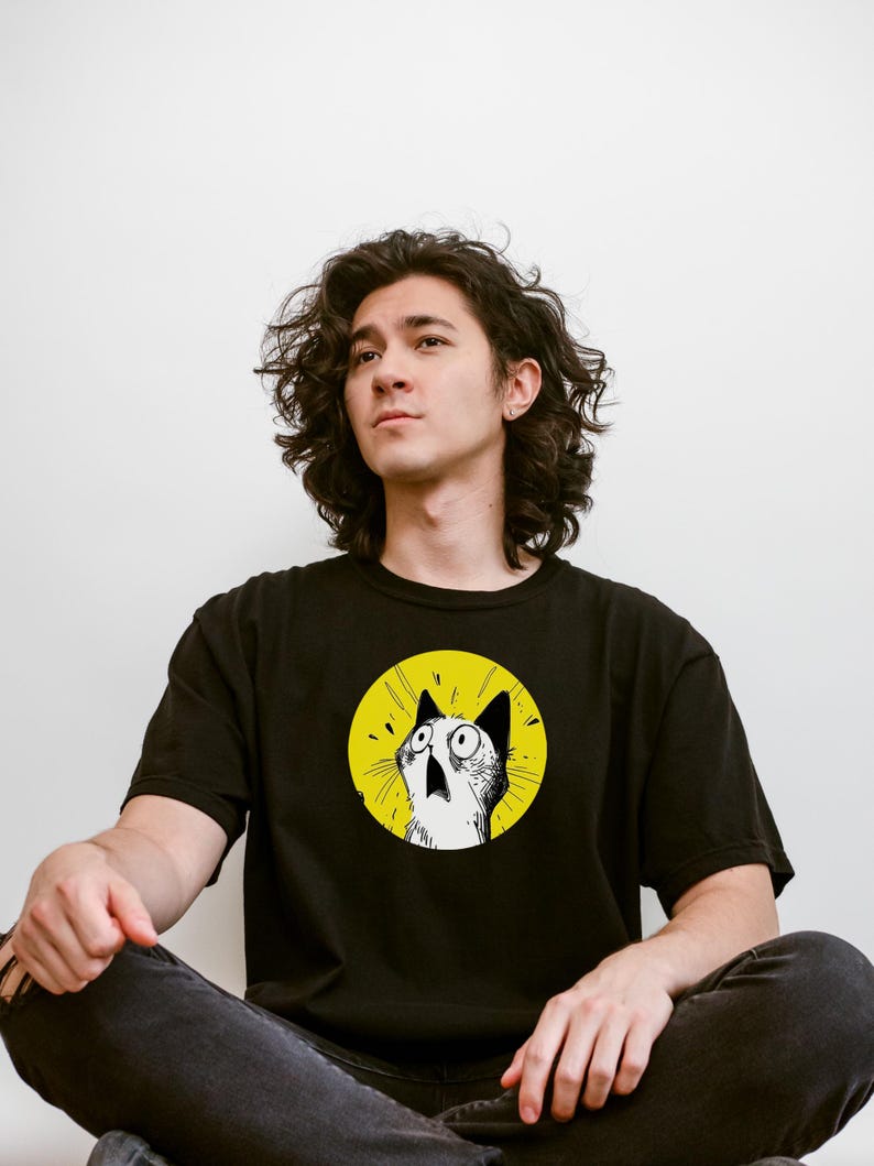 May include: A black short-sleeved t-shirt featuring a circular graphic on the chest. The graphic depicts a white cat with wide, surprised eyes and an open mouth, set against a bright yellow background with black radiating lines.