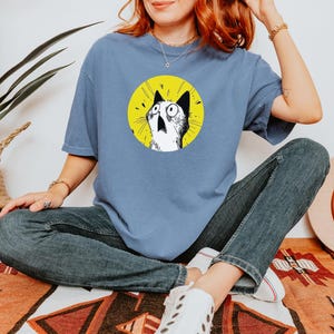 May include: A dusty blue short-sleeve t-shirt featuring a circular graphic on the chest. The graphic depicts a black and white illustration of a cat with wide, surprised eyes against a bright yellow background. The shirt has a crew neckline.