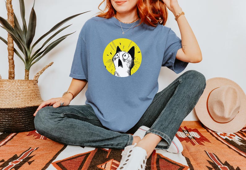 May include: A dusty blue short-sleeve t-shirt featuring a circular graphic on the chest. The graphic depicts a black and white illustration of a cat with wide, surprised eyes against a bright yellow background. The shirt has a crew neckline.